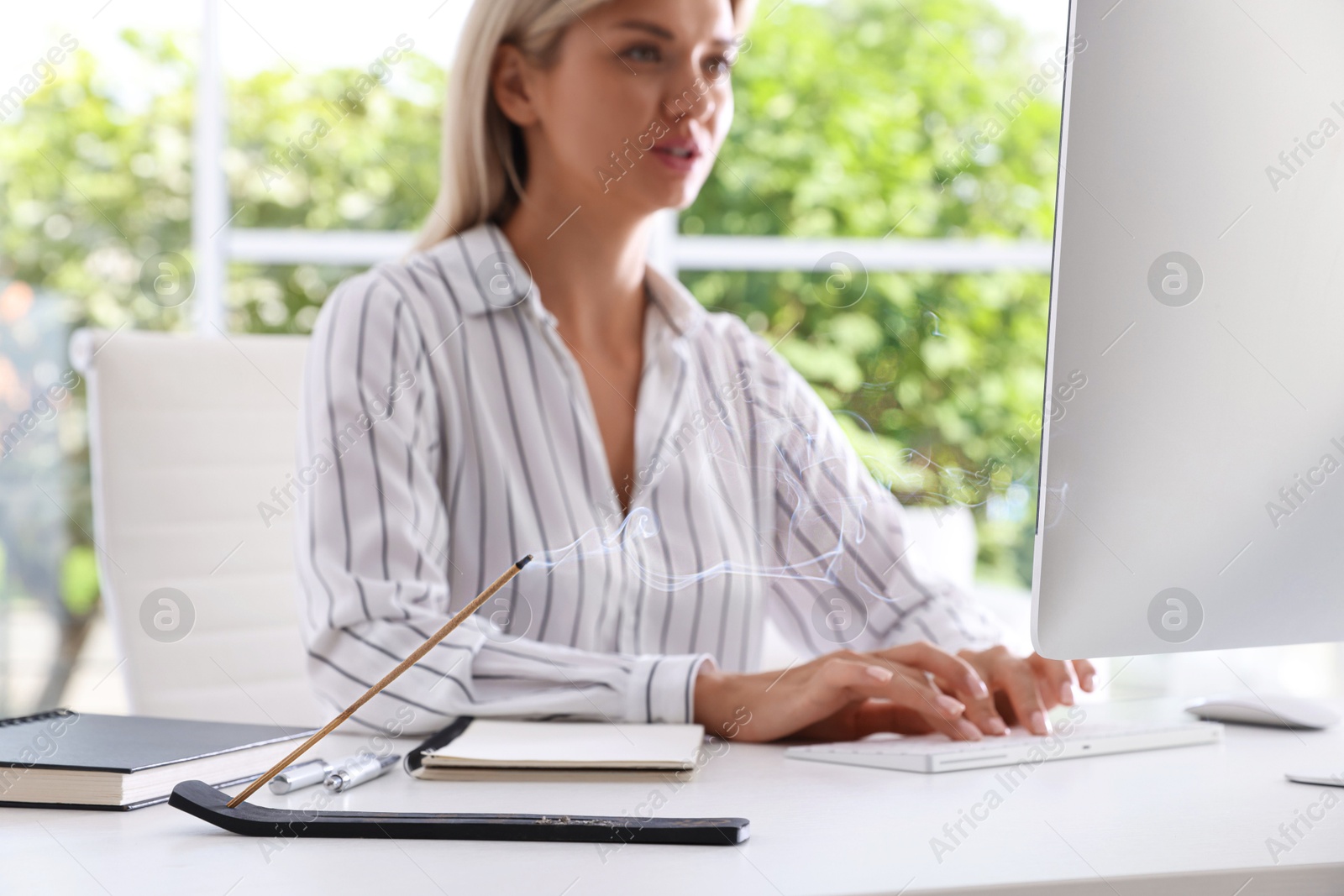 Photo of Woman working with computer at table while incense stick smoldering in holder indoors, selective focus