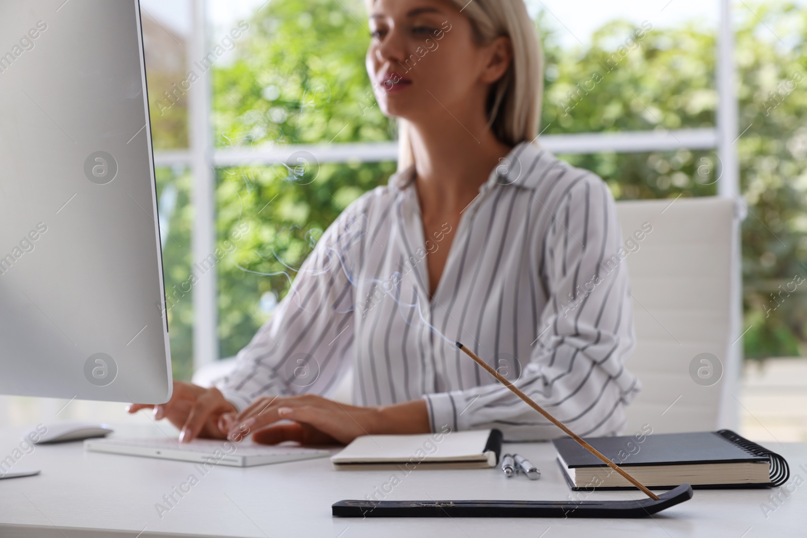 Photo of Woman working with computer at table while incense stick smoldering in holder indoors, selective focus