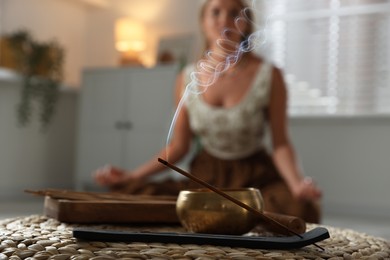 Photo of Young woman practicing yoga indoors, focus on smoldering incense stick and tibetan singing bowl