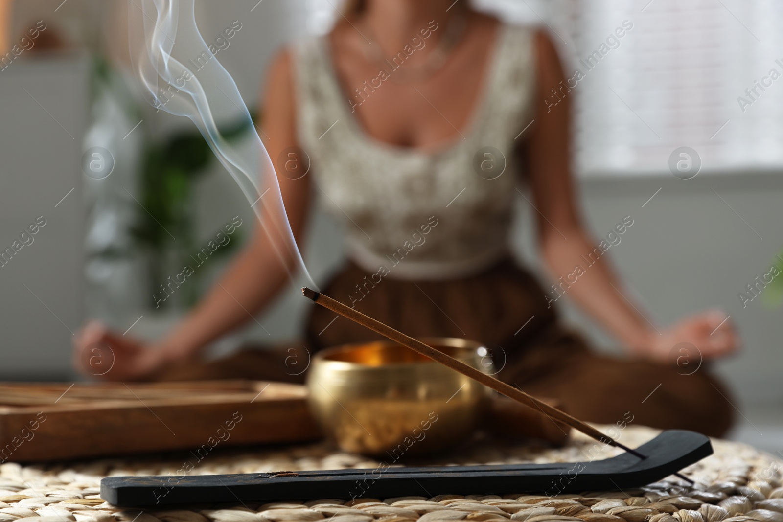 Photo of Young woman practicing yoga indoors, focus on smoldering incense stick and tibetan singing bowl