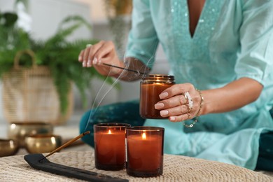 Photo of Woman lighting incense sticks with burning candles indoors, closeup