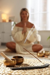 Photo of Young woman practicing yoga on floor indoors, focus on smoldering incense stick and tibetan singing bowls