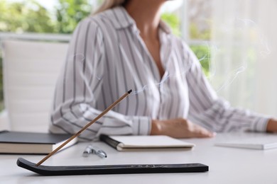 Photo of Young woman working at table while incense stick smoldering in holder indoors, selective focus