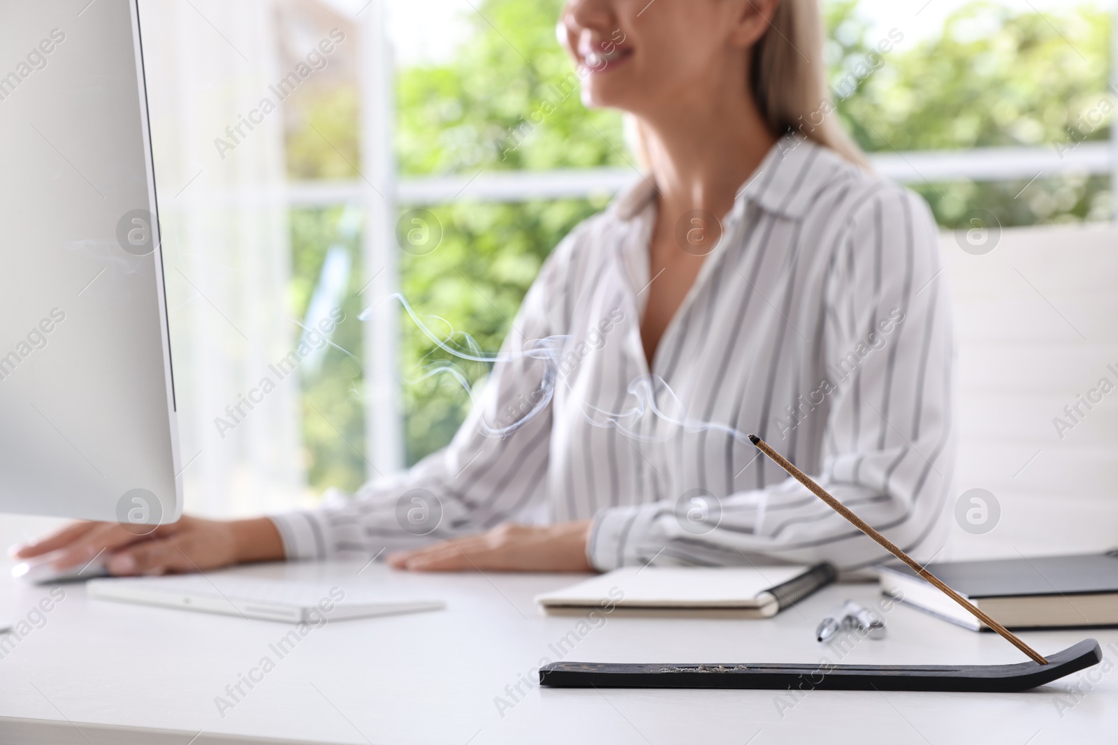 Photo of Young woman working at table while incense stick smoldering in holder indoors, selective focus