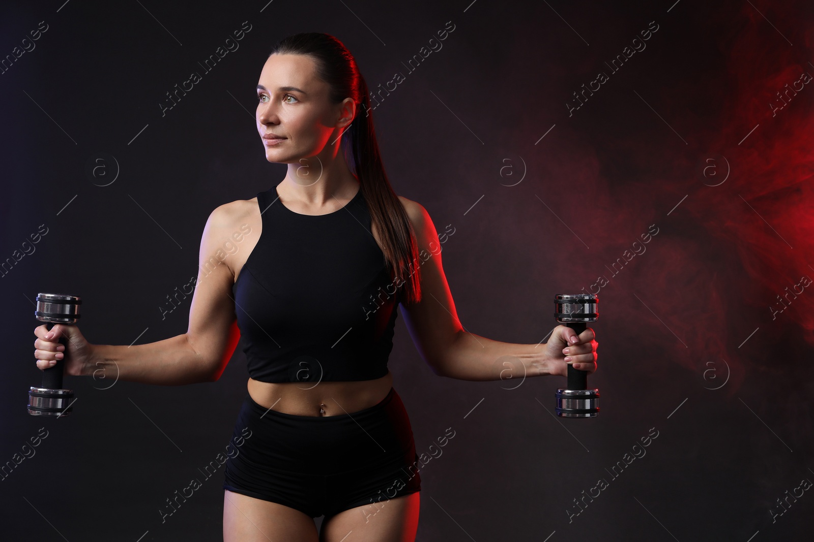 Photo of Woman exercising with dumbbells in smoke on dark background. Space for text