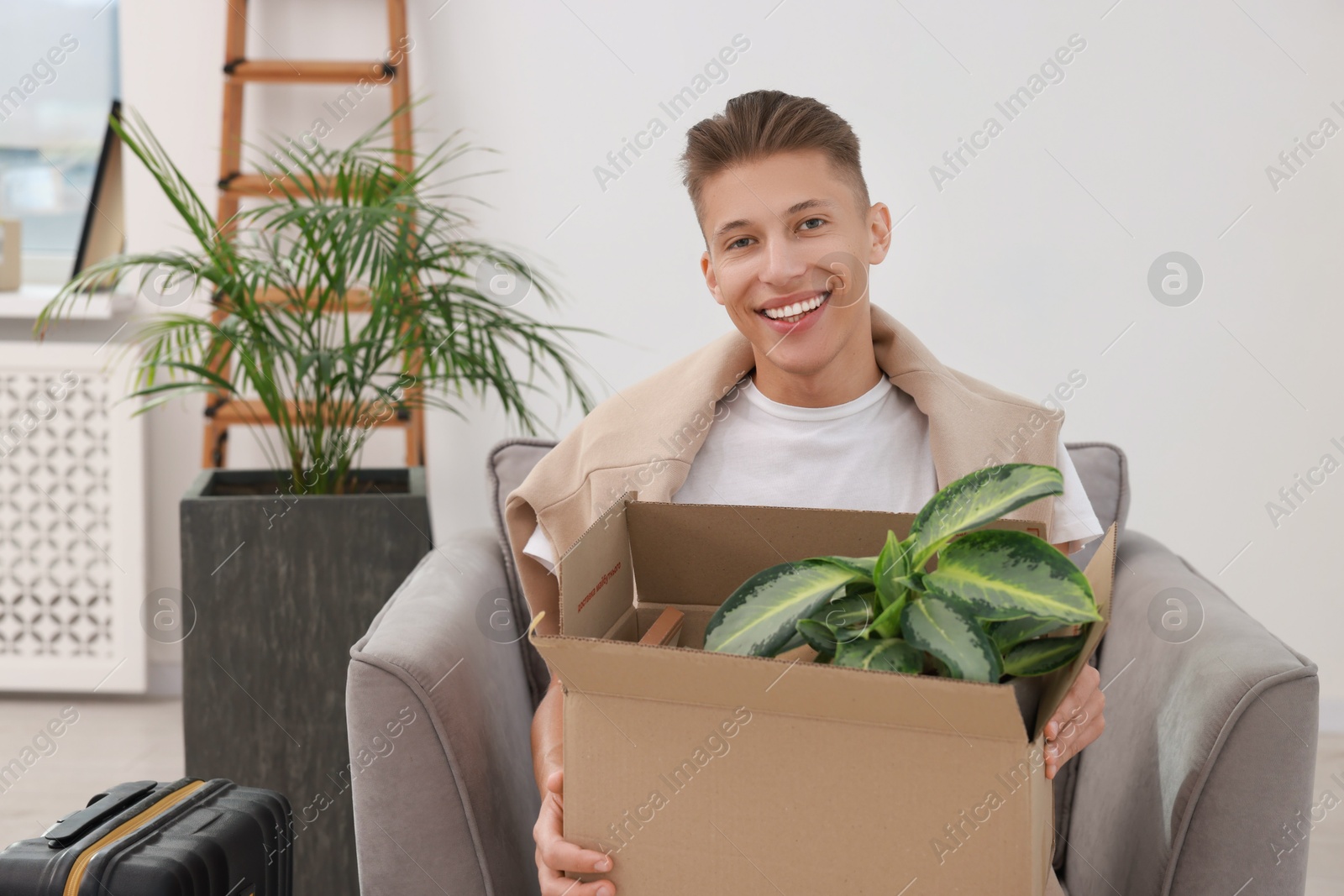 Photo of Happy man holding moving box with houseplant in new apartment. Housewarming party