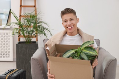 Photo of Happy man holding moving box with houseplant in new apartment. Housewarming party