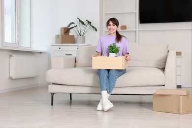 Photo of Happy woman holding wooden crate with stuff on sofa in new apartment. Housewarming party