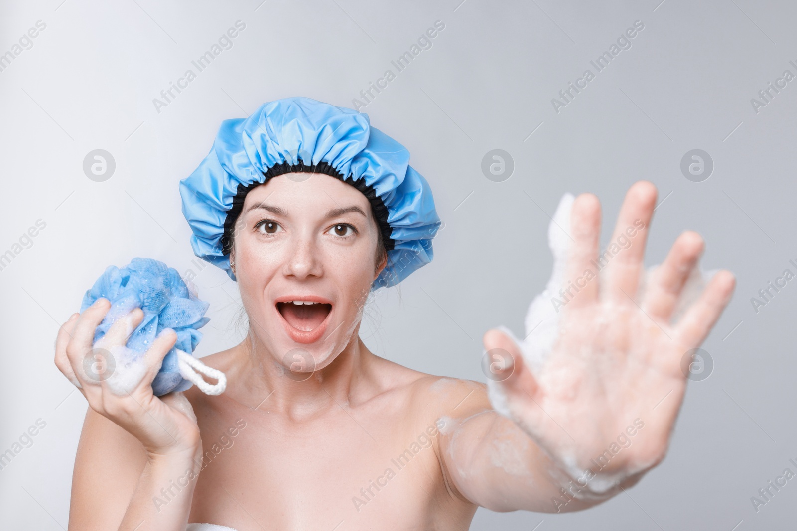 Photo of Emotional woman with shower cap and mesh sponge making stop gesture on grey background