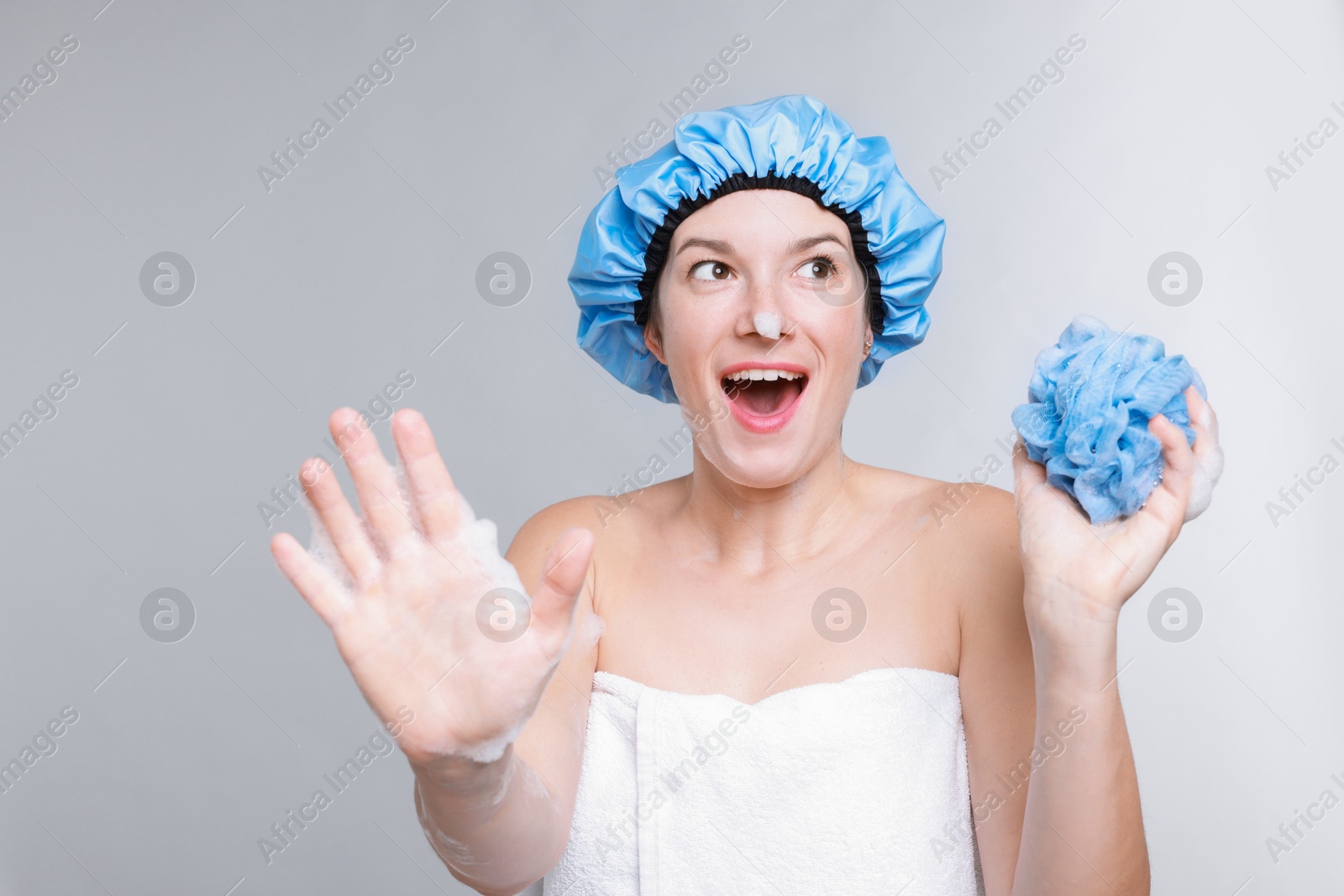 Photo of Emotional woman with shower cap and mesh sponge making stop gesture on grey background