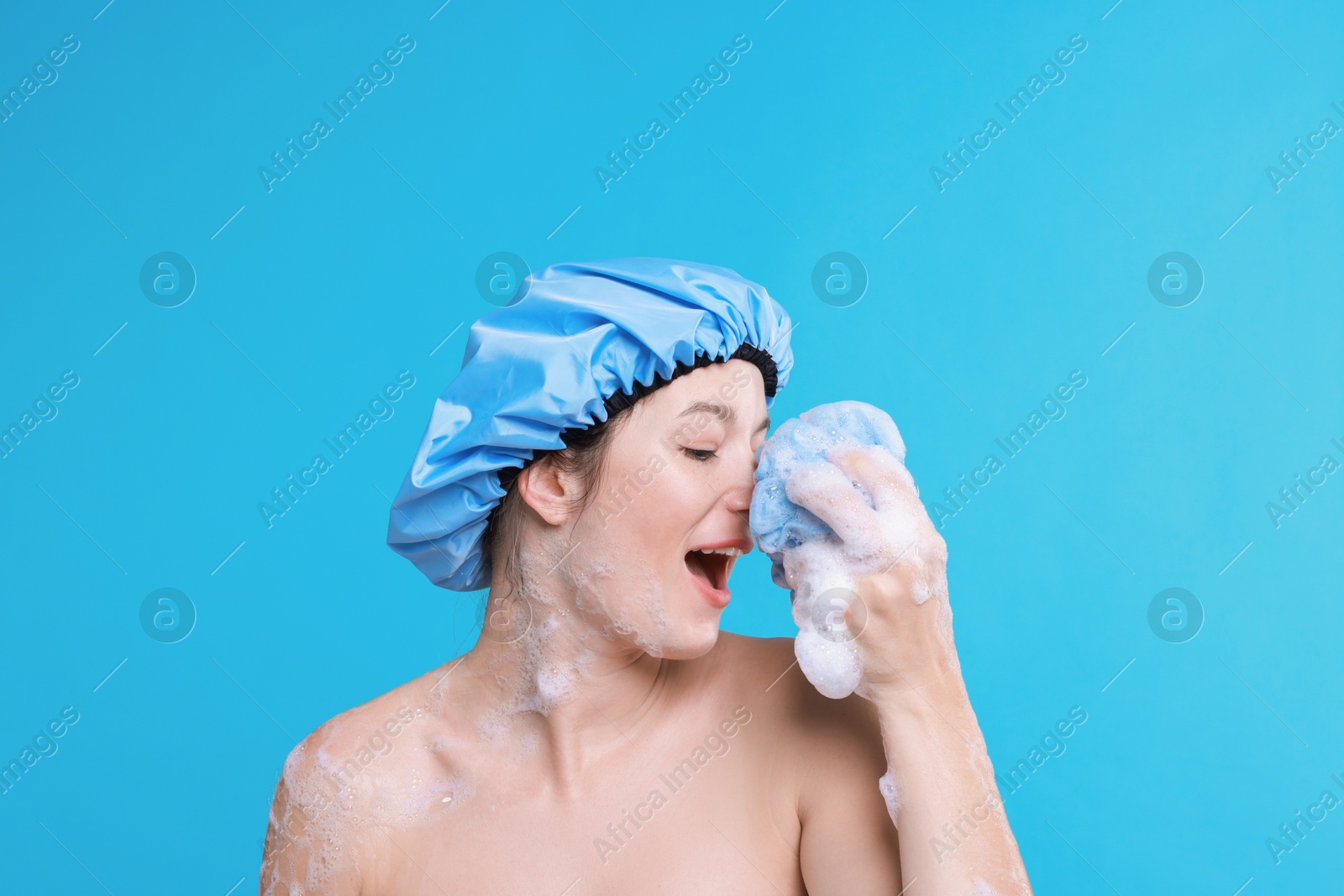 Photo of Woman in shower cap covered with soap foam holding mesh sponge on light blue background