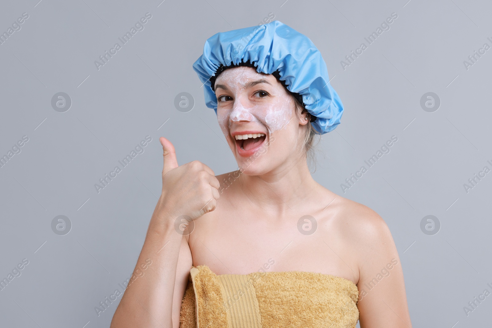 Photo of Woman with shower cap and cream on her face showing thumbs up against grey background