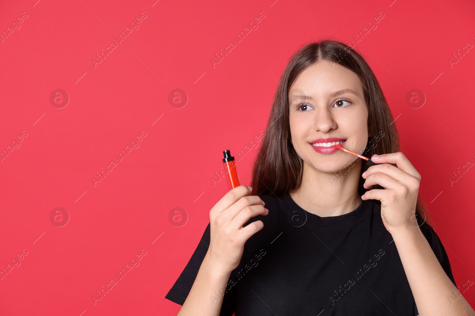 Photo of Smiling teenage girl applying lip gloss on red background. Space for text