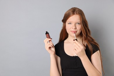 Teenage girl applying lip gloss on grey background. Space for text