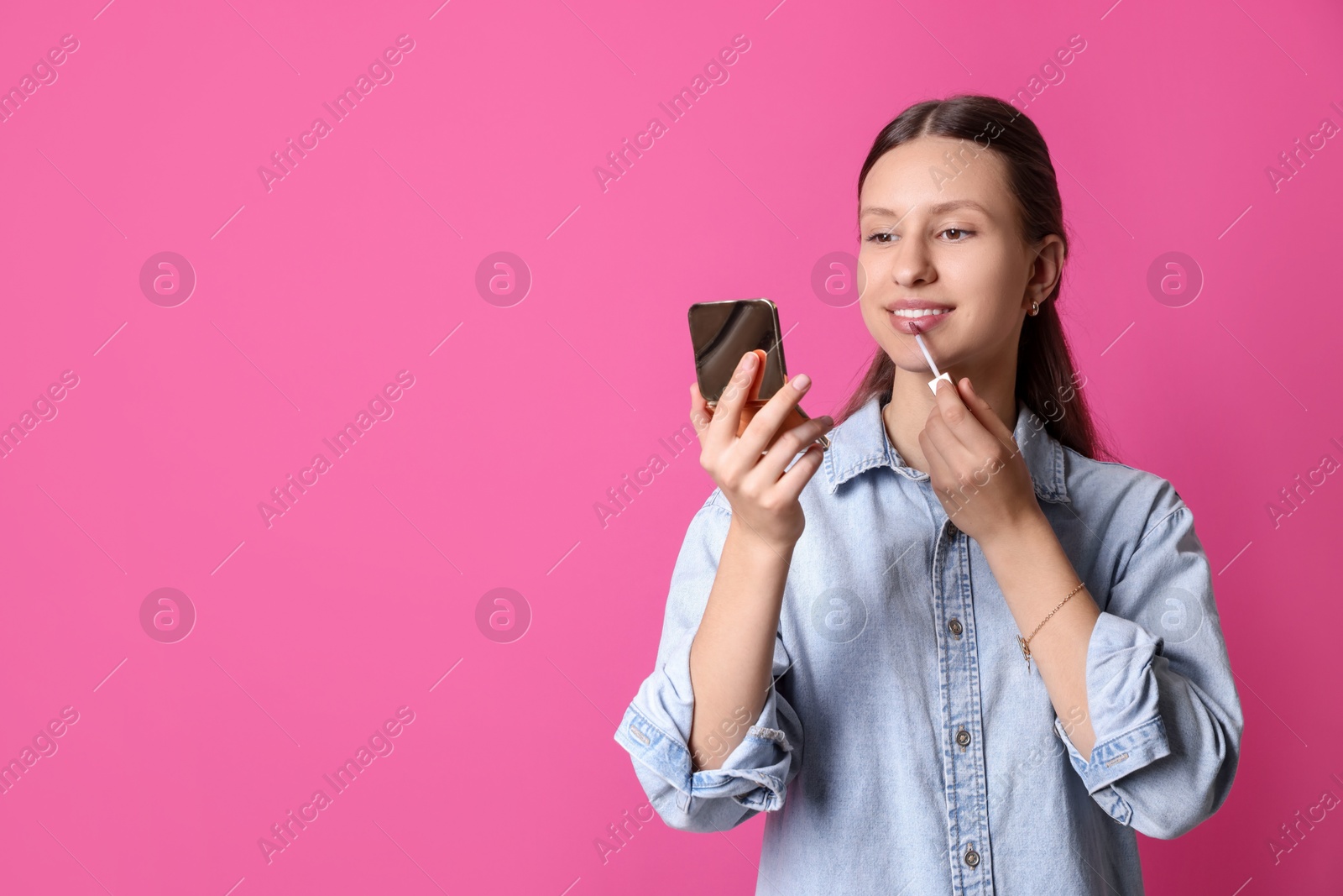 Photo of Smiling teenage girl applying lip gloss on pink background. Space for text