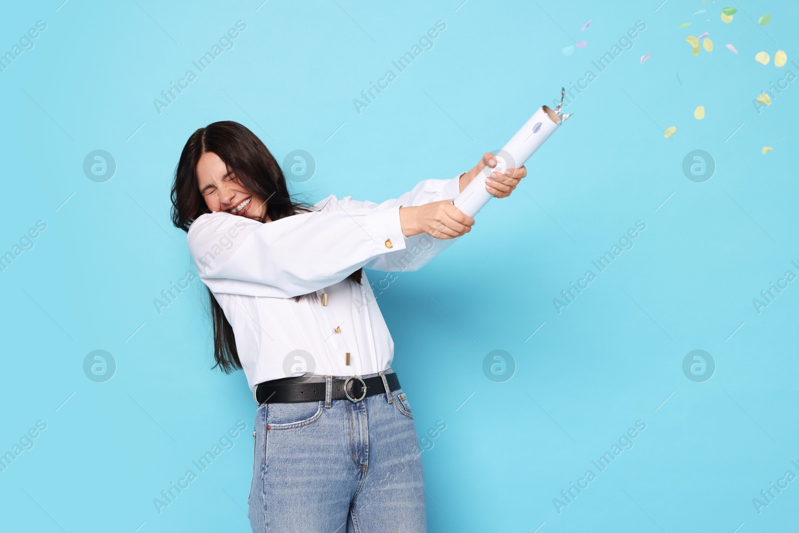 Photo of Happy woman blowing up confetti popper on light blue background. Surprise party