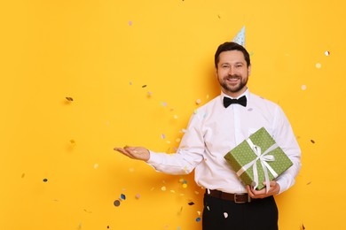 Photo of Happy man with gift box under falling confetti on orange background, space for text. Surprise party