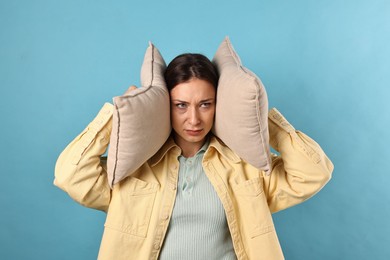 Annoyed woman covering her ears with pillows due to loud sound on light blue background