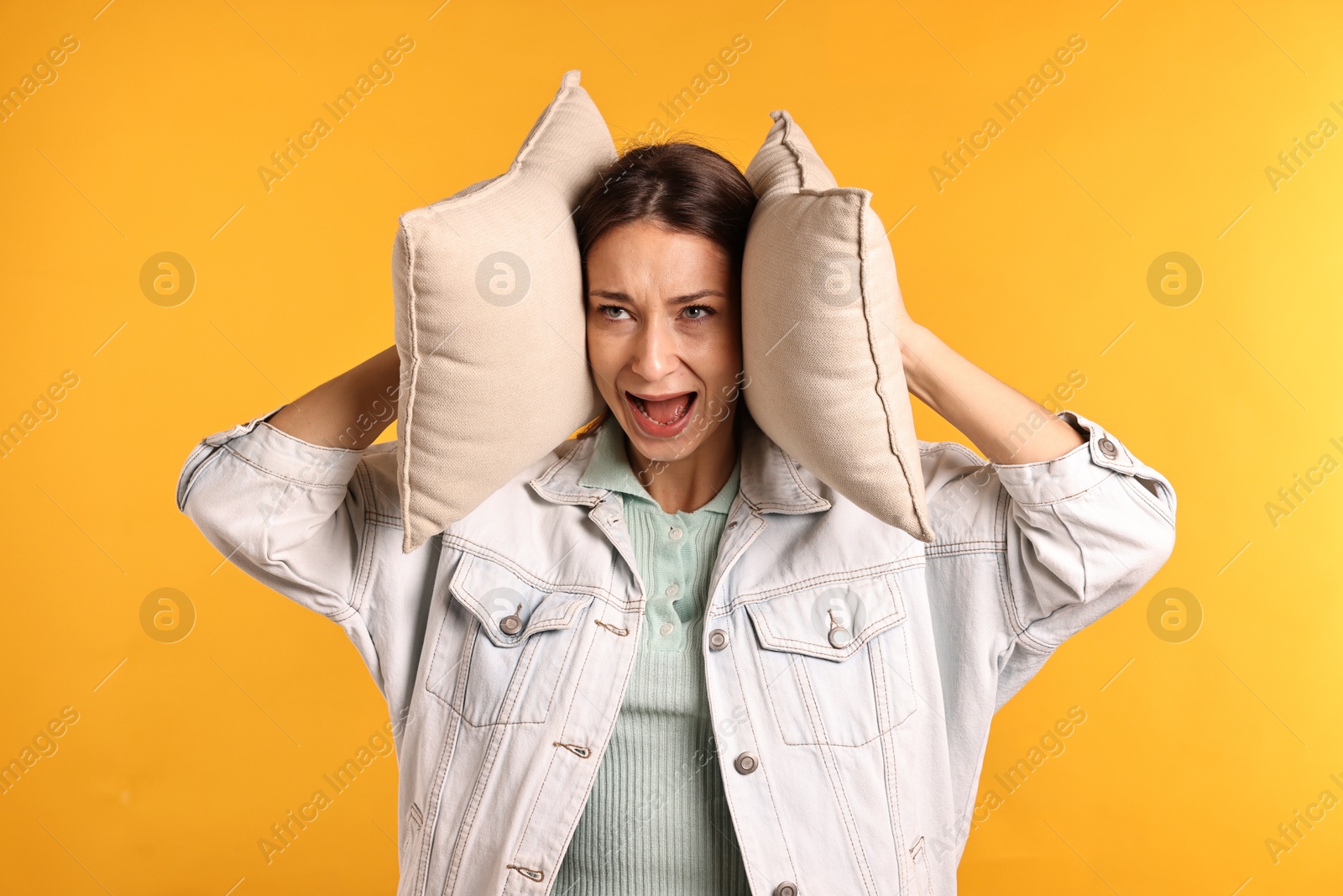 Photo of Annoyed woman covering her ears with pillows due to loud sound on orange background