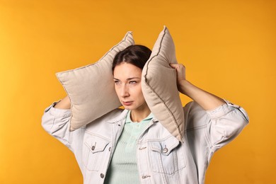 Photo of Annoyed woman covering her ears with pillows due to loud sound on orange background