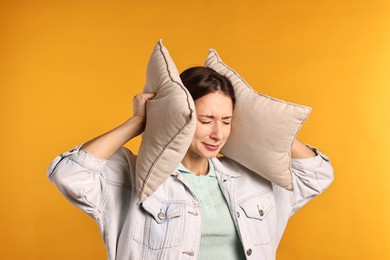 Photo of Annoyed woman covering her ears with pillows due to loud sound on orange background