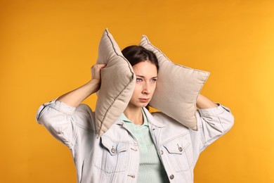 Annoyed woman covering her ears with pillows due to loud sound on orange background