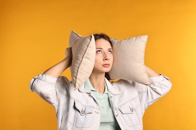 Photo of Annoyed woman covering her ears with pillows due to loud sound on orange background