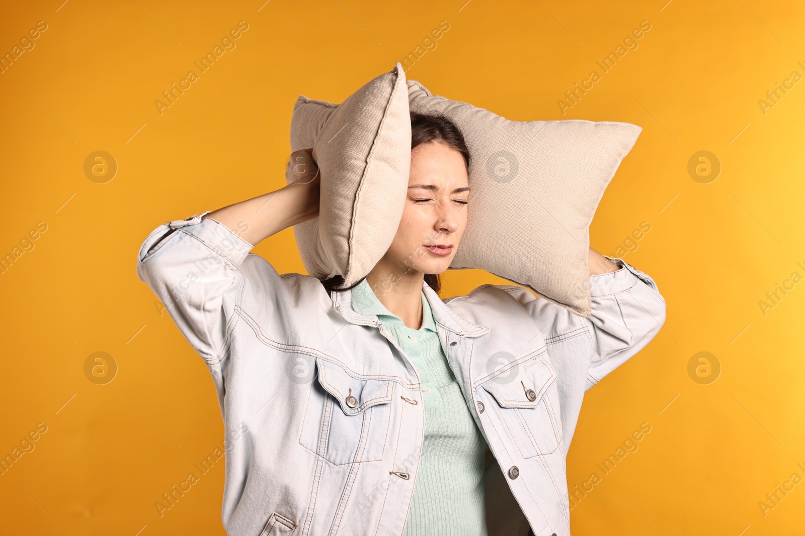 Photo of Annoyed woman covering her ears with pillows due to loud sound on orange background