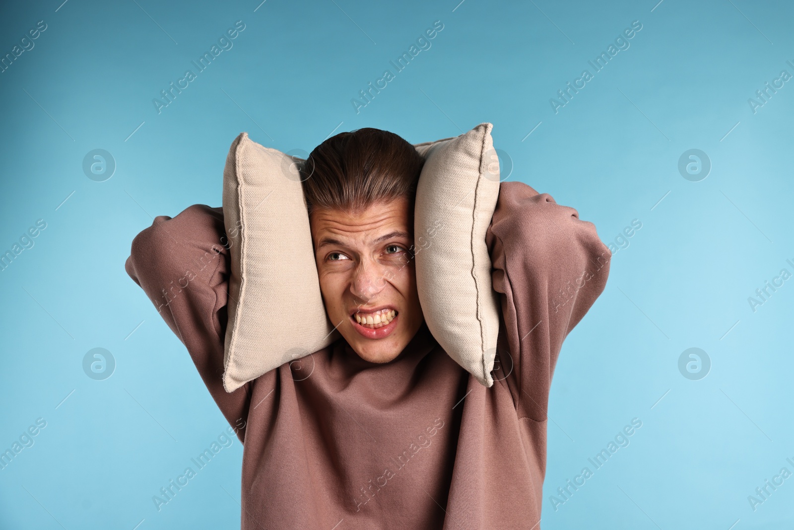 Photo of Annoyed young man covering his ears with pillows due to loud sound on light blue background