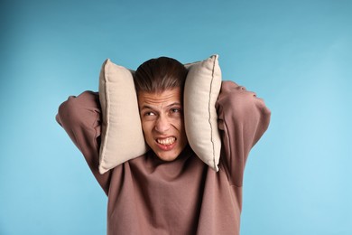 Annoyed young man covering his ears with pillows due to loud sound on light blue background