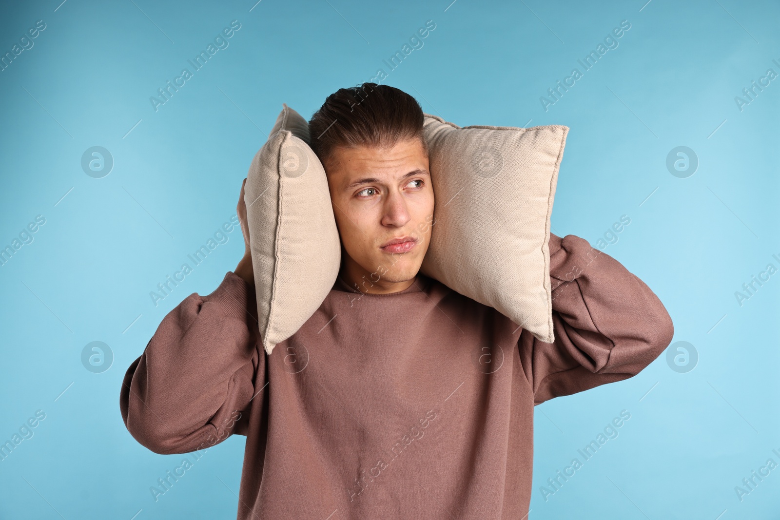 Photo of Annoyed young man covering his ears with pillows due to loud sound on light blue background