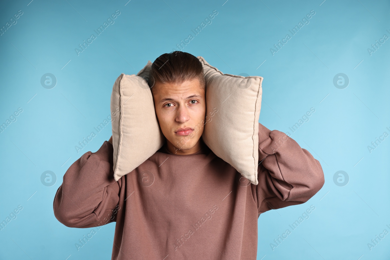 Photo of Annoyed young man covering his ears with pillows due to loud sound on light blue background