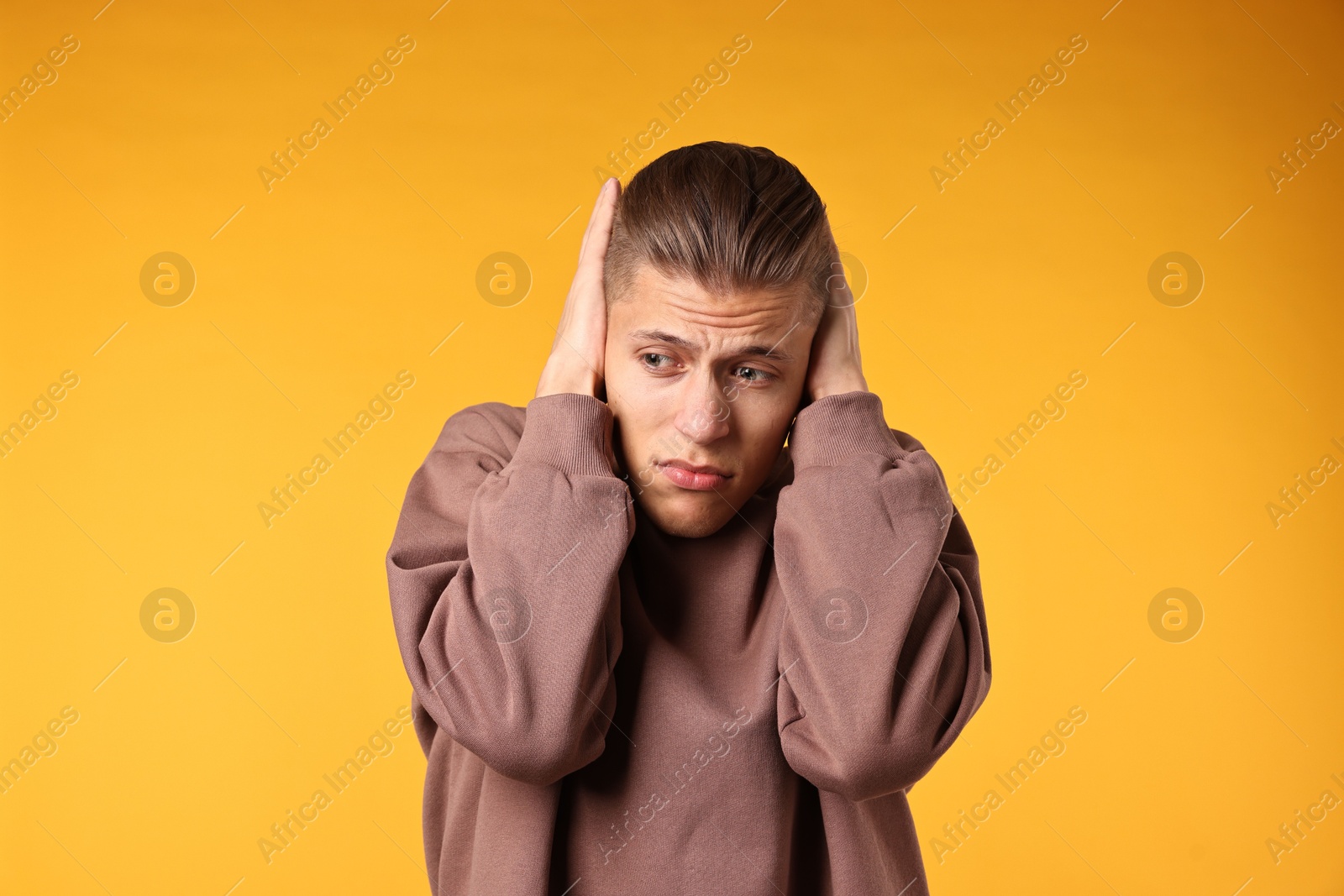 Photo of Annoyed young man covering his ears due to loud sound on orange background