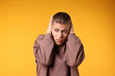 Photo of Annoyed young man covering his ears due to loud sound on orange background