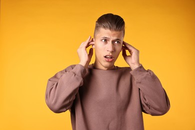 Photo of Annoyed young man covering his ears due to loud sound on orange background