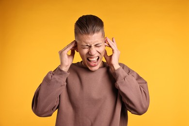 Photo of Annoyed young man covering his ears due to loud sound on orange background