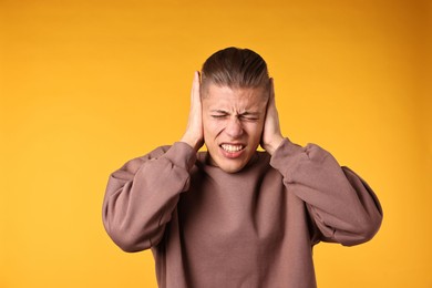 Photo of Annoyed young man covering his ears due to loud sound on orange background