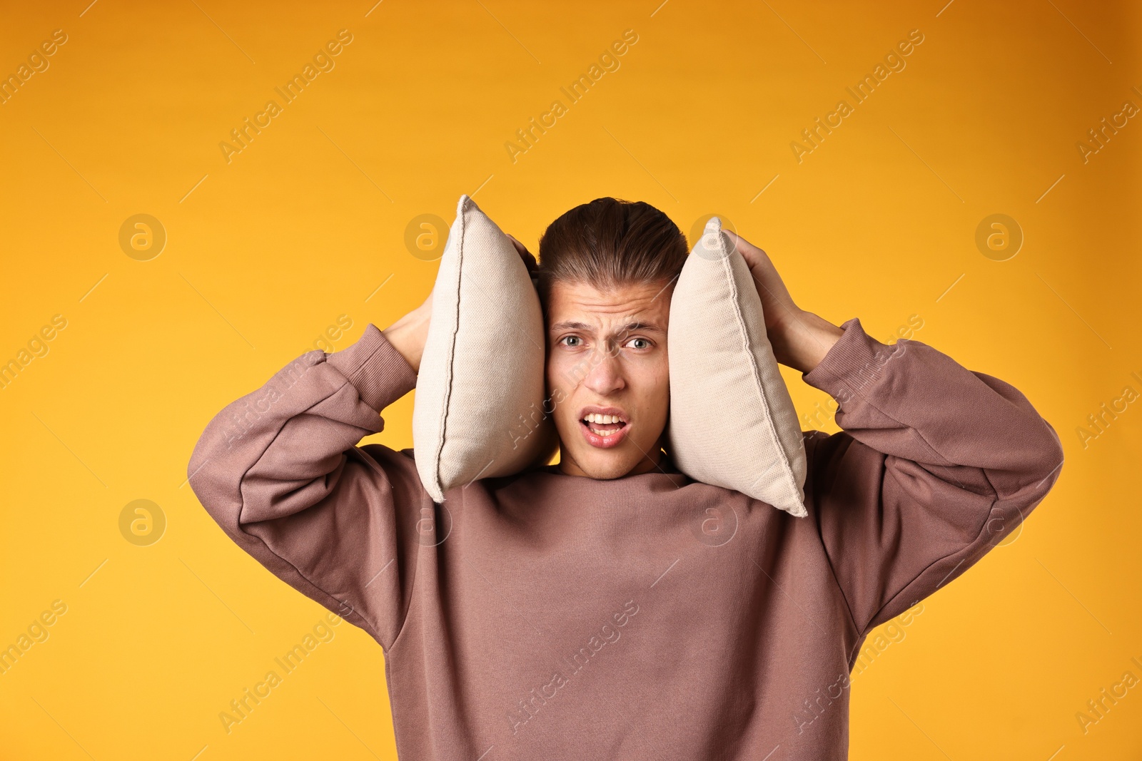 Photo of Annoyed young man covering his ears with pillows due to loud sound on orange background