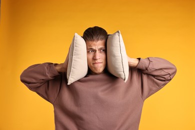 Photo of Annoyed young man covering his ears with pillows due to loud sound on orange background