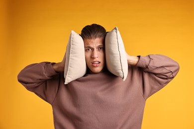 Photo of Annoyed young man covering his ears with pillows due to loud sound on orange background