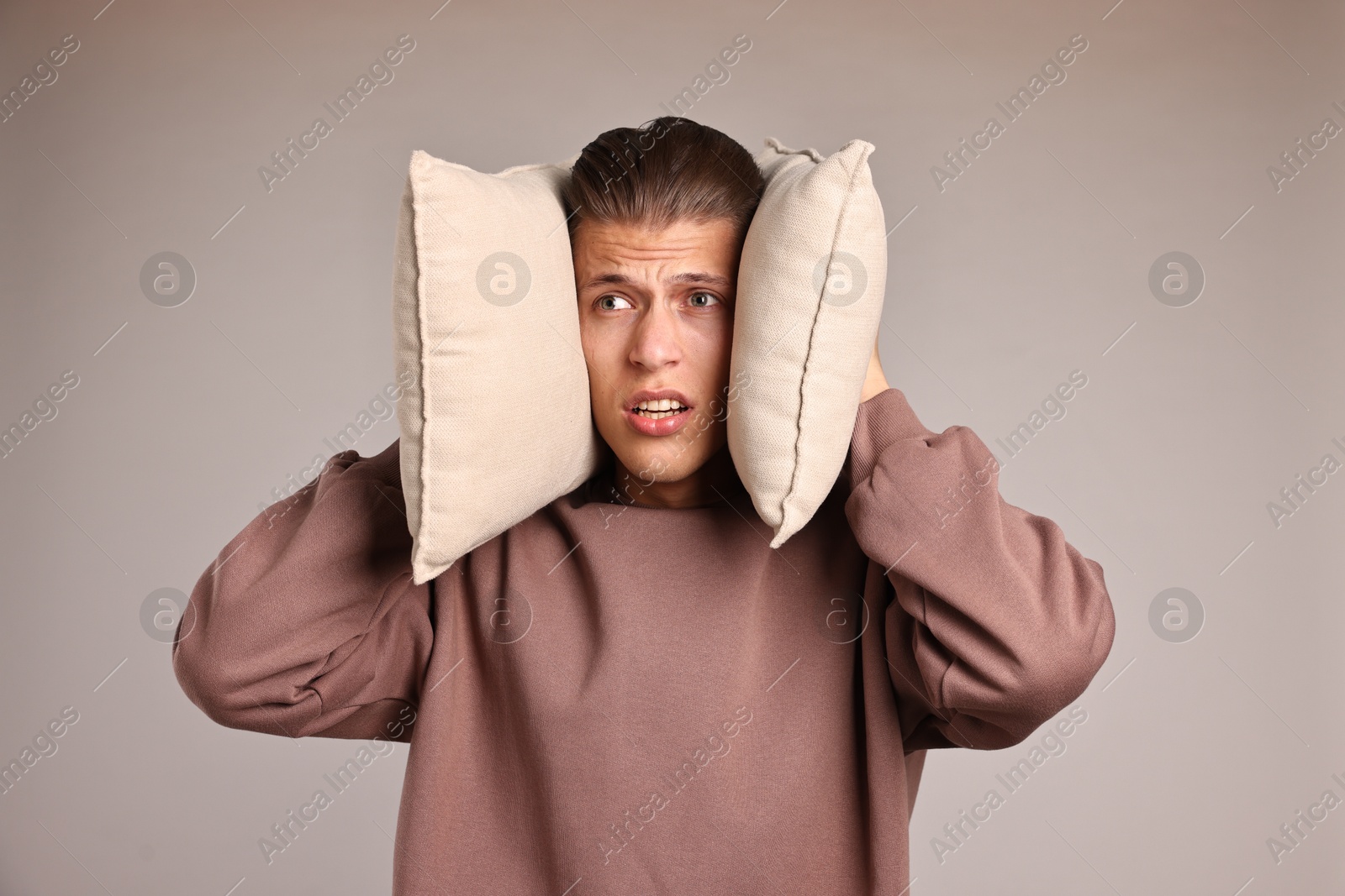 Photo of Annoyed young man covering his ears with pillows due to loud sound on light grey background