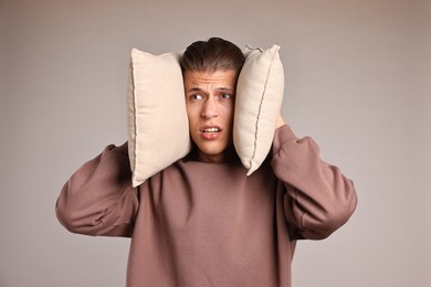 Annoyed young man covering his ears with pillows due to loud sound on light grey background