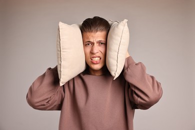 Annoyed young man covering his ears with pillows due to loud sound on light grey background