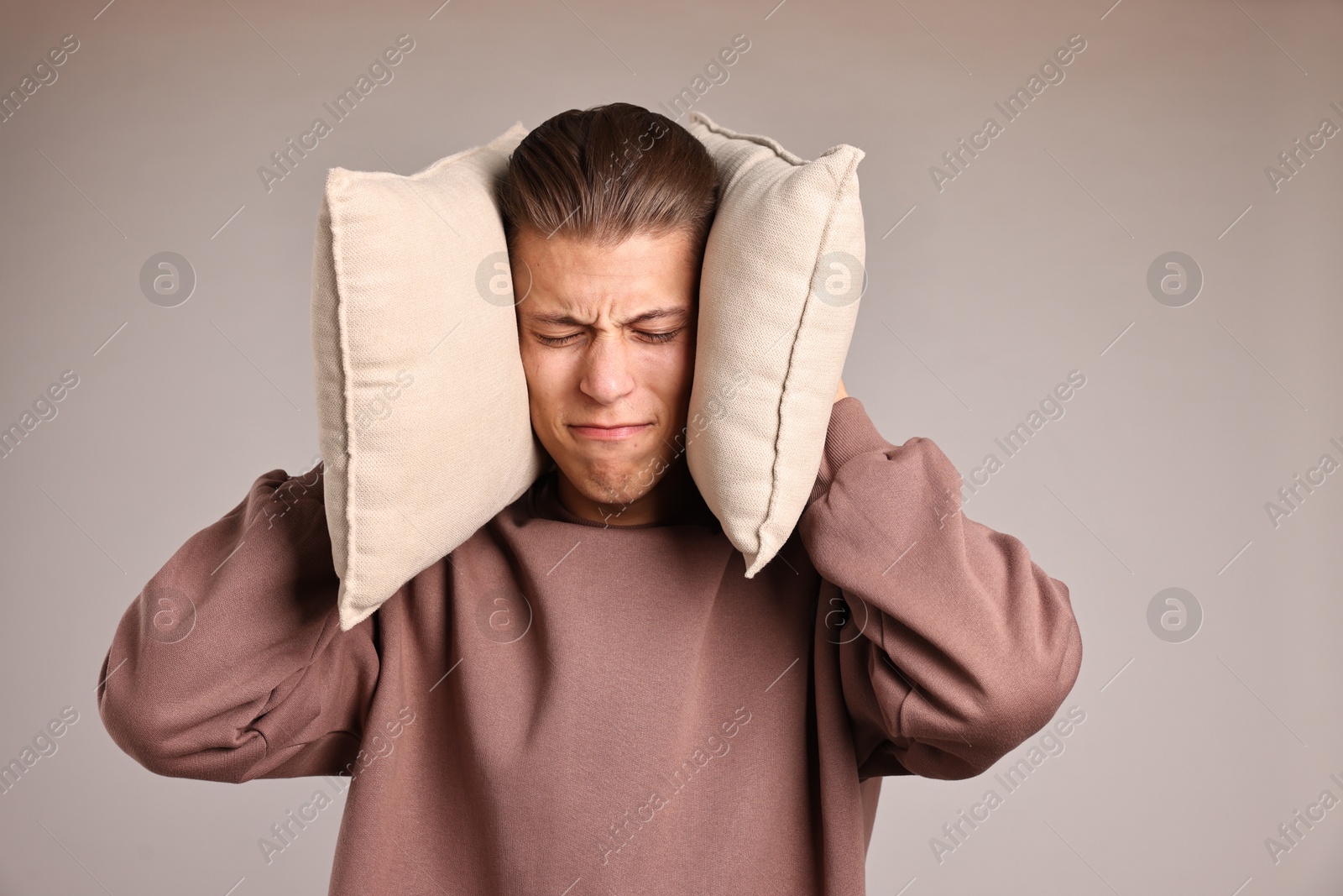 Photo of Annoyed young man covering his ears with pillows due to loud sound on light grey background