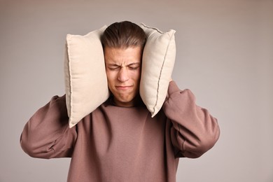 Annoyed young man covering his ears with pillows due to loud sound on light grey background