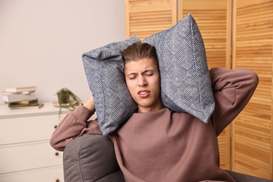 Annoyed young man covering his ears with pillows due to loud sound at home