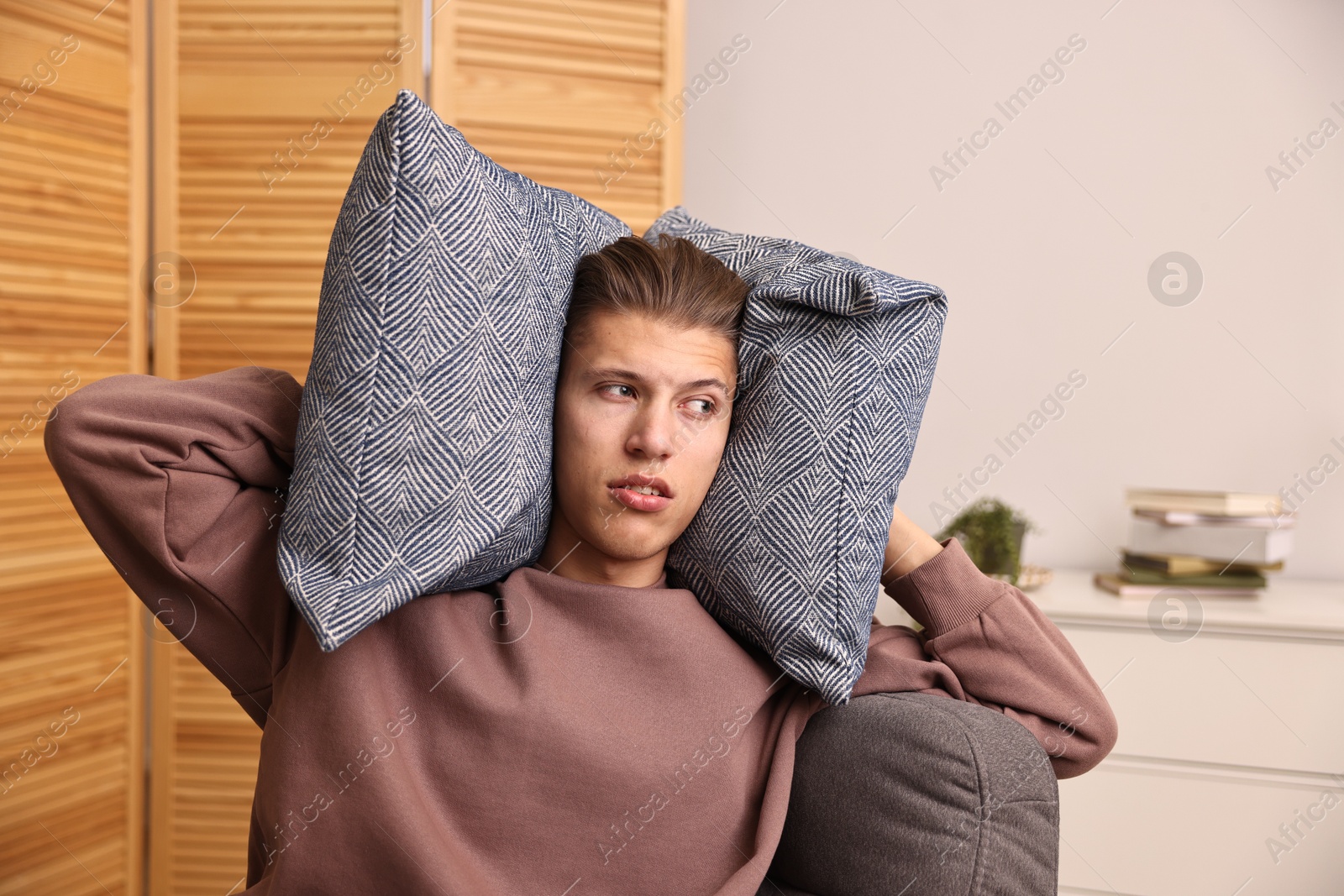 Photo of Annoyed young man covering his ears with pillows due to loud sound at home