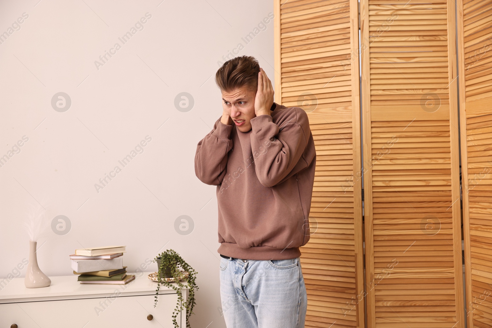 Photo of Annoyed young man covering his ears due to loud sound at home