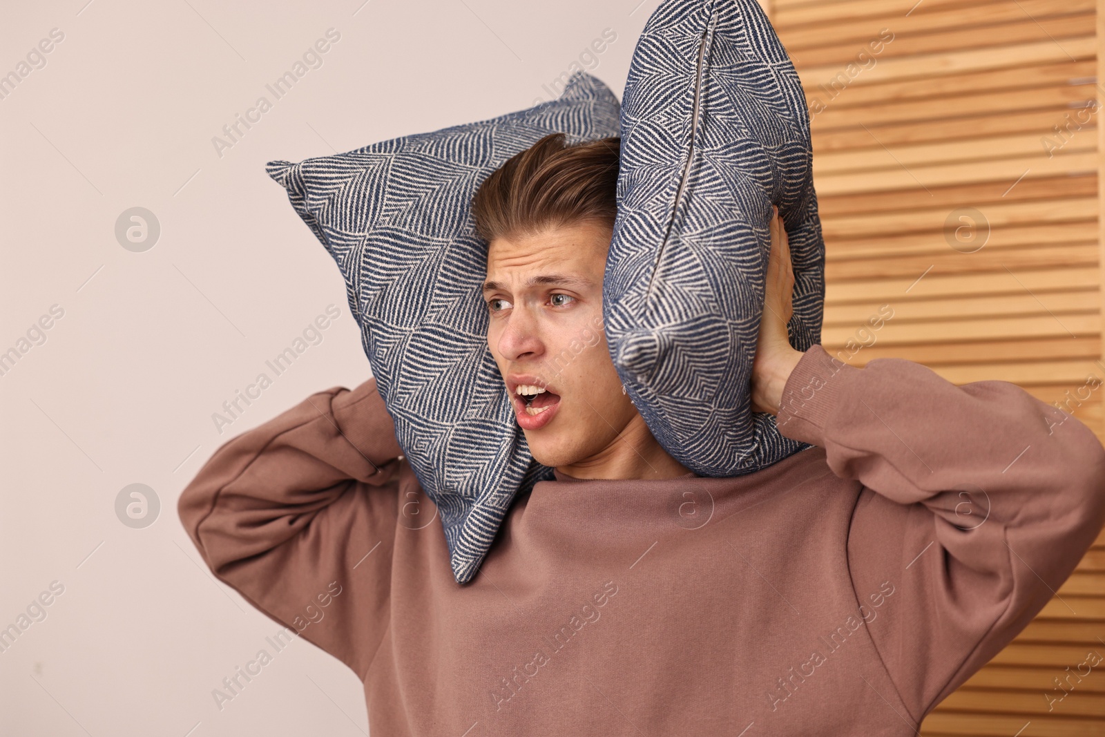 Photo of Annoyed young man covering his ears with pillows due to loud sound at home