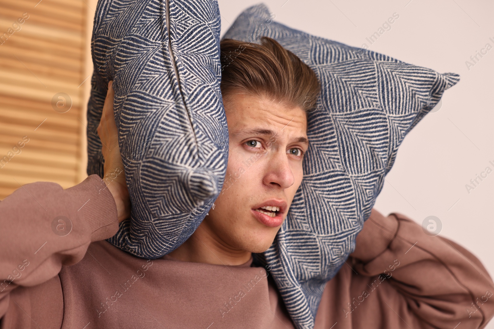 Photo of Annoyed young man covering his ears with pillows due to loud sound at home